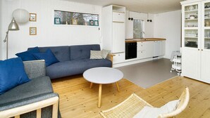 Ett kompakt vardagsrum med en soffa, ett litet runt bord, en fåtölj i rotting, en liten köksdel och ett skåp med glasdörrar.