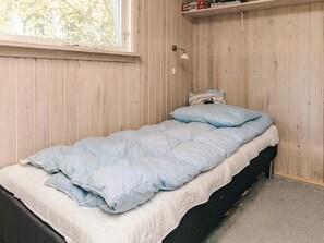 A single bed with a blue blanket and pillows, a bedside table with a lamp, and a shelf with books and a small item. - 5 Person Holiday Home in Hurup Thy (Hurup Thy)