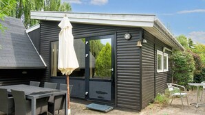 A small, dark-colored building with a black door and a white umbrella. There is a table and chairs outside, and a bell on the wall. - 6 Person Holiday Home in Follenslev (Follenslev)