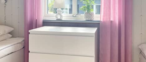 A room with a white chest of drawers, a bedside table with a lamp, and a window with pink curtains.
