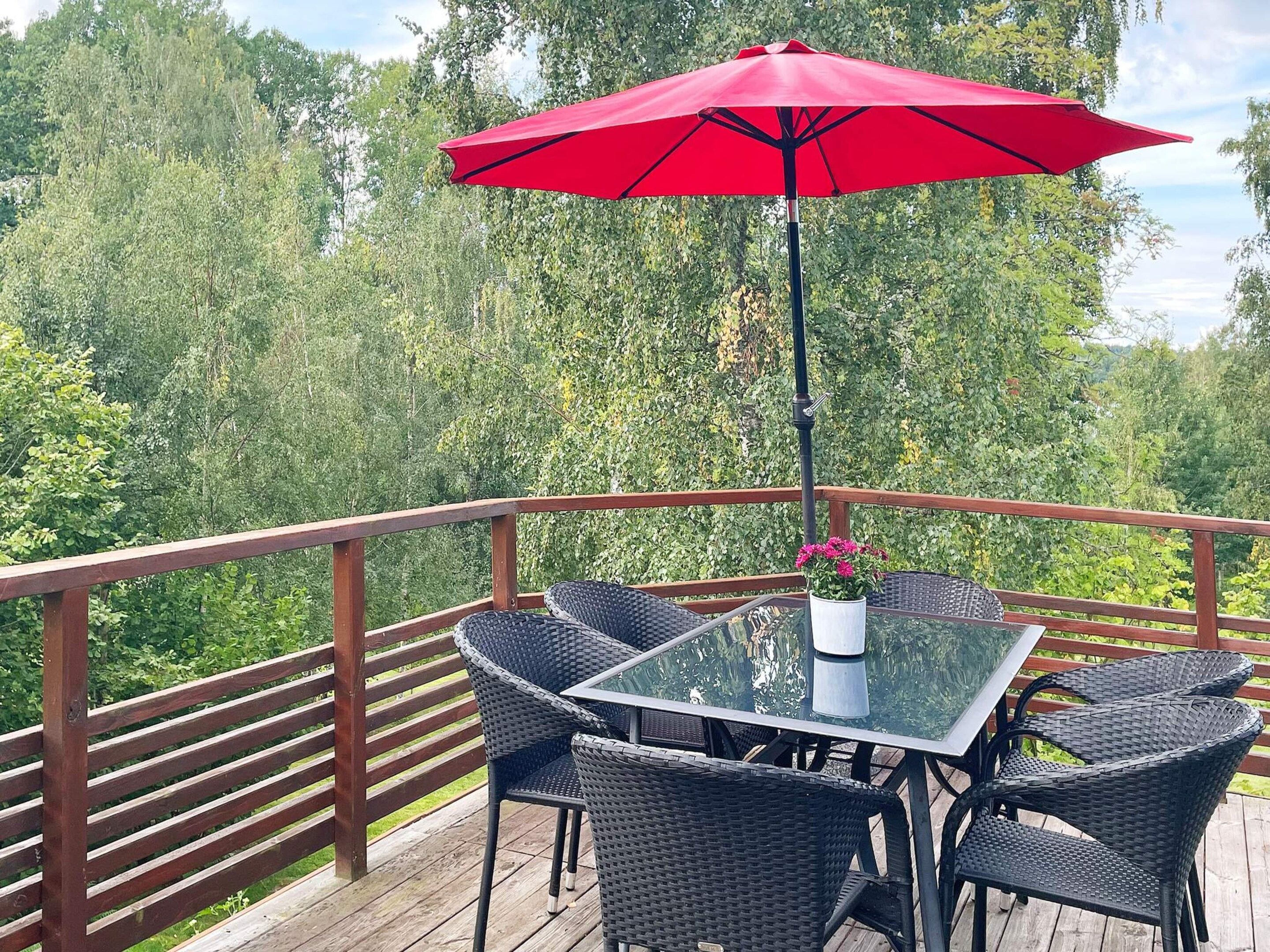 Mesa con cubierta de vidrio, cuatro sillas de mimbre y una sombrilla roja sobre una terraza de madera con barandilla.