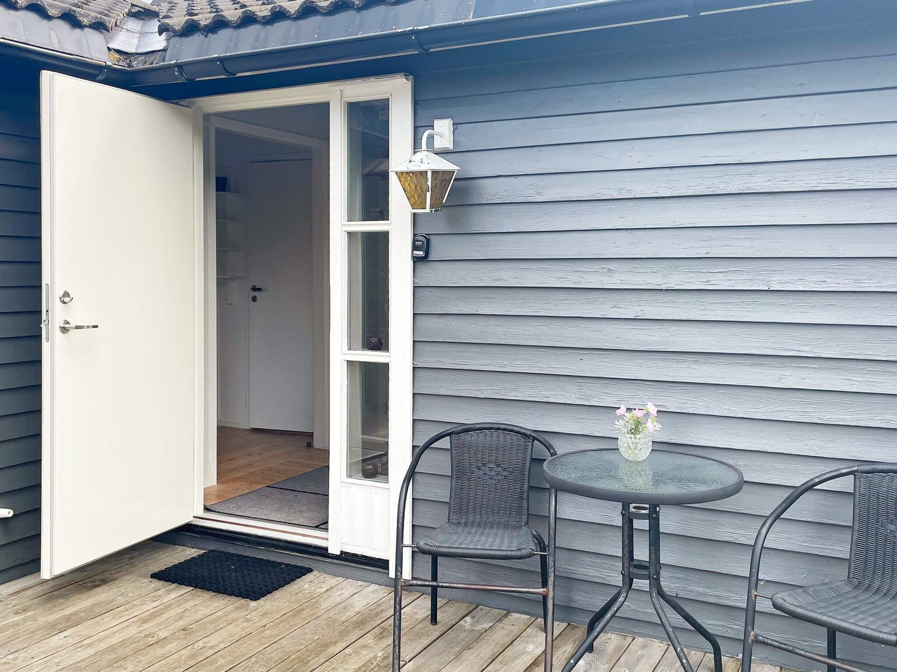 A wooden deck with a white door leading to an interior room, a round table with chairs, and a wall-mounted light fixture.