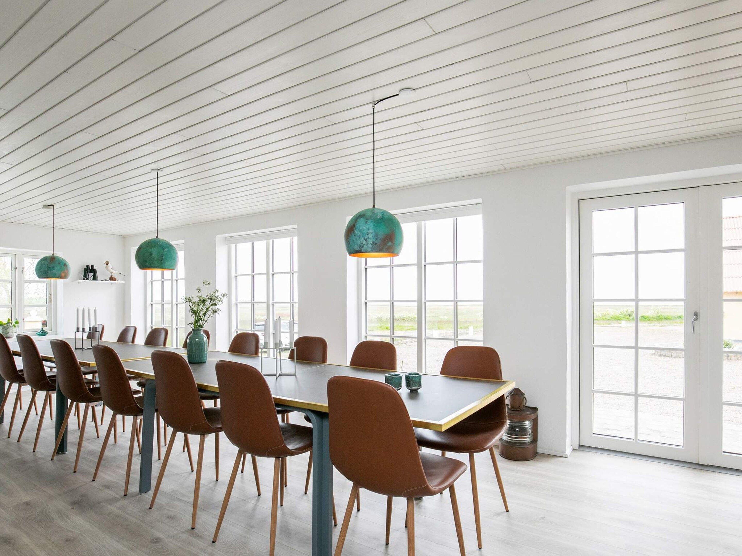 Un comedor amplio con una larga mesa, varias sillas, lámparas colgantes y ventanas grandes.