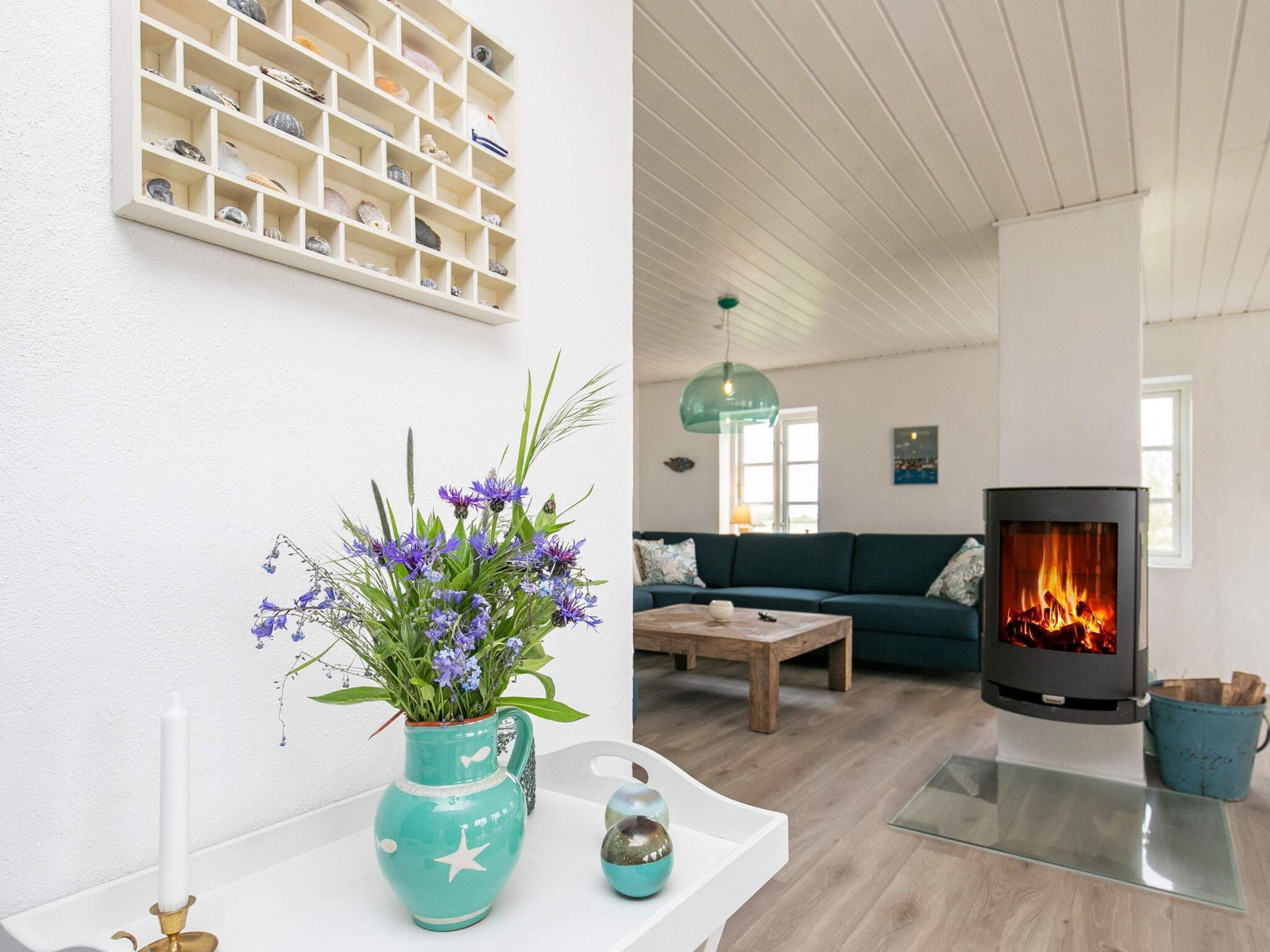 A living room with a fireplace, a wall-mounted shelf with decorative items, and a teal vase with flowers.