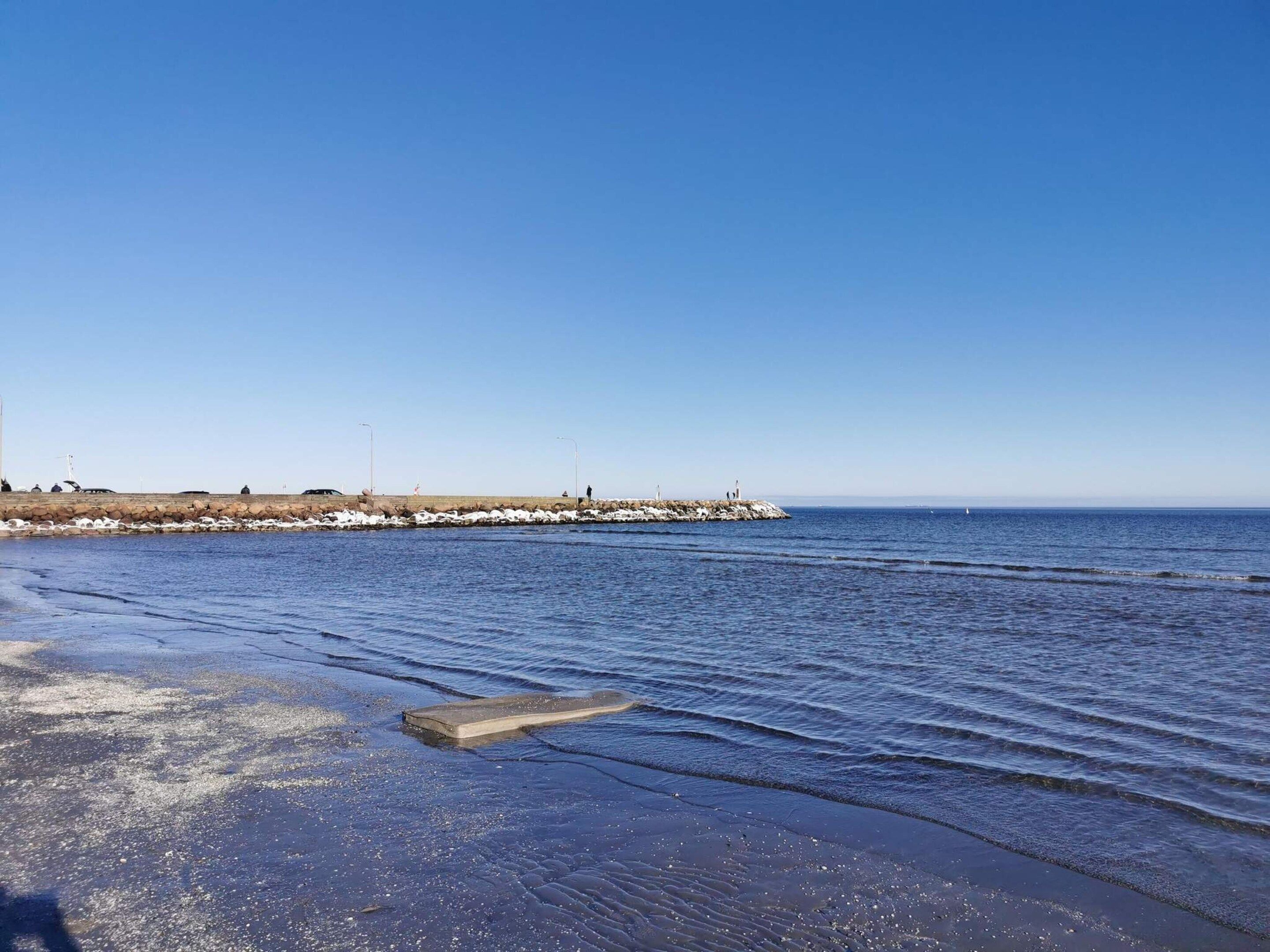Paesaggio costiero con un molo in cemento che si protende nell'acqua, cielo sereno e mare calmo.