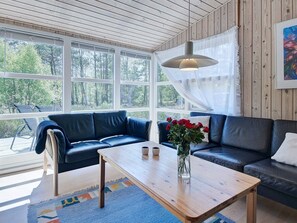 A cozy living room with a wooden ceiling, large windows, a blue sofa, a wooden coffee table, and a vase with red flowers. - 4 Person Holiday Home in Nexo (Nexø)
