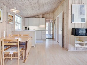 A modern interior with a wooden floor, a dining area with a table and chairs, a kitchen with white cabinets, and a TV on a shelf. - 4 Person Holiday Home in Nexo (Nexø)