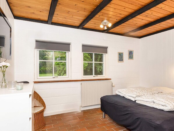 Et soveværelse med en seng, to vinduer, en radiator, træloft og en vægmonteret lampe.