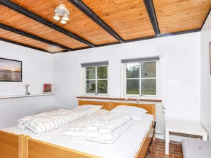 A bedroom with a wooden bed, two windows, a white brick wall, and a small bedside table.