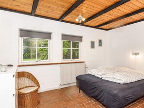 A room with a bed, a chair, two windows, a radiator, and a ceiling with wooden beams. - 6 Person Holiday Home in Bindslev (Bindslev)