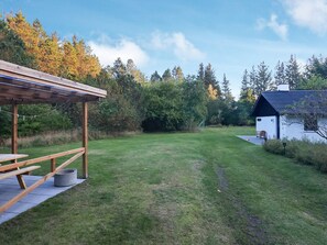 Et græsklædt udendørsareal med en træpergola og en hvid bygning med mørkt tag.
