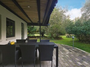 An outdoor patio with a table and chairs, a covered area, and a view of a garden with trees and a lawn.