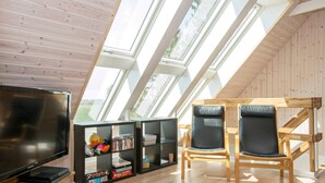 A room with a sloped ceiling, skylights, a flat-screen TV, two black leather chairs, and wooden shelves with books and games. - 10 Person Holiday Home in Glesborg (Glesborg)