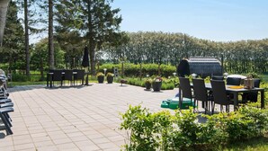An outdoor patio with tiled flooring, multiple tables and chairs, a barbecue grill, and surrounding greenery. - 10 Person Holiday Home in Glesborg (Glesborg)