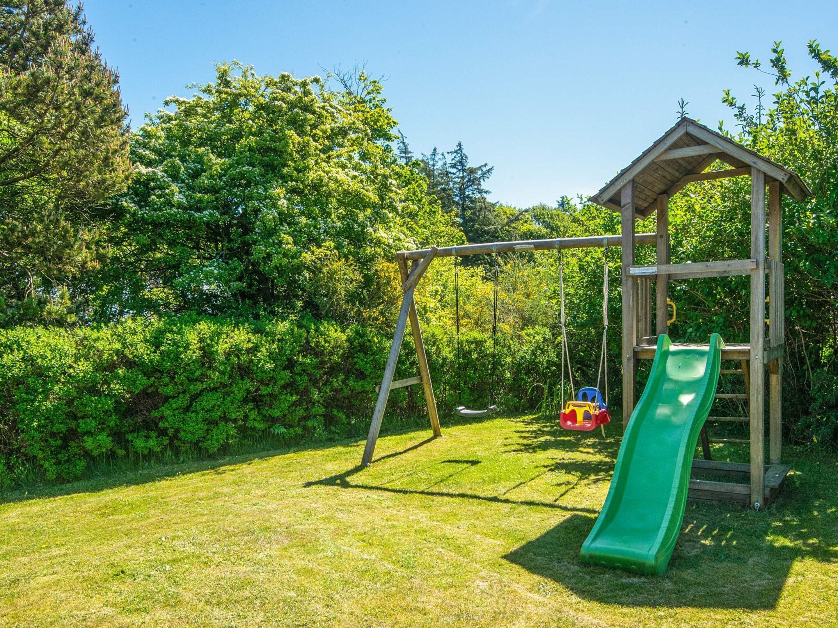 A wooden play structure with a slide and swings in a grassy area surrounded by trees.