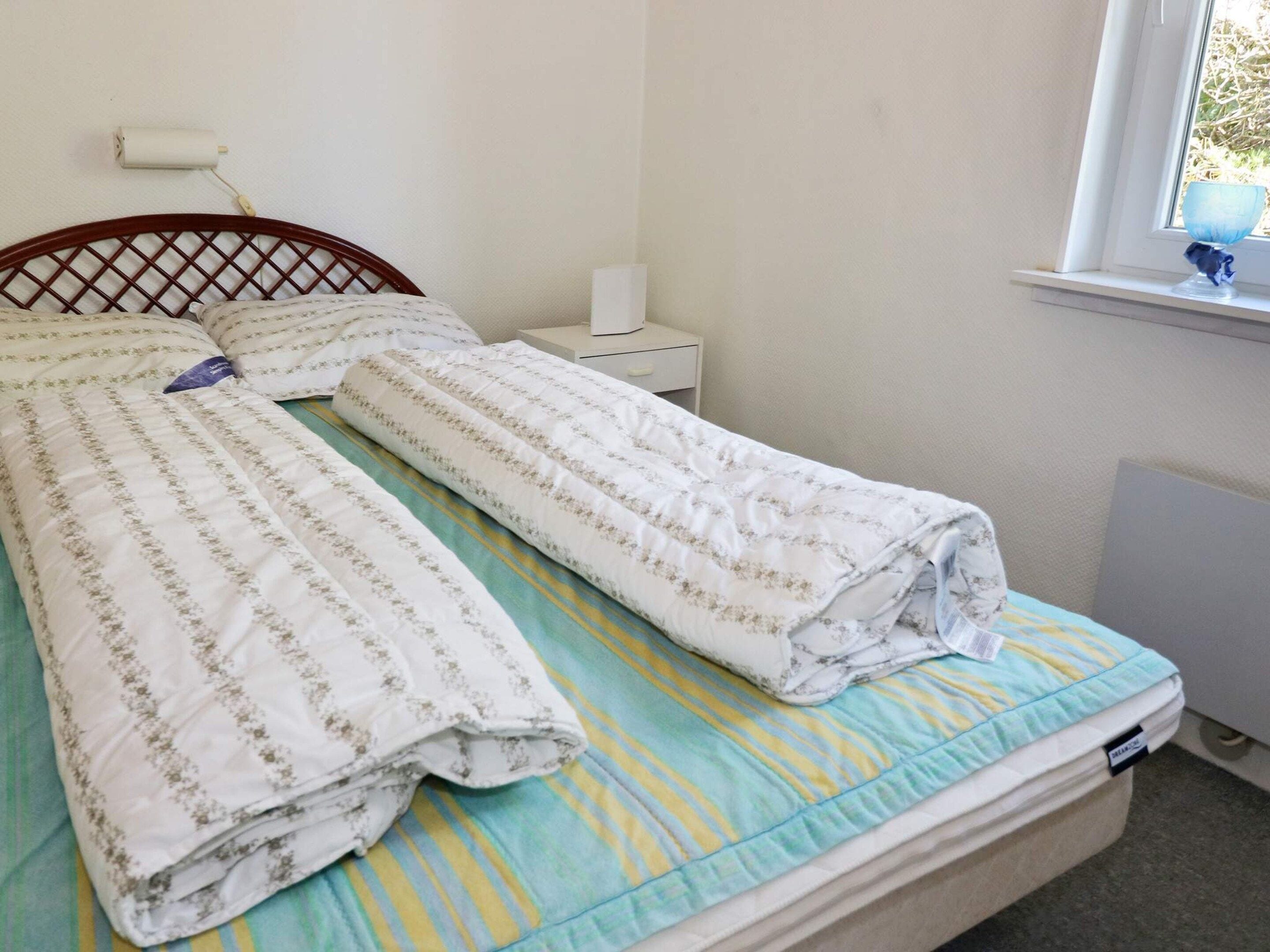 A bed with a striped bedspread and two pillows, a bedside table with a lamp, and a window with a view of greenery.