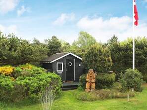 Een kleine hut met een zwarte deur en een raam, omgeven door groen en een vlaggenmast.