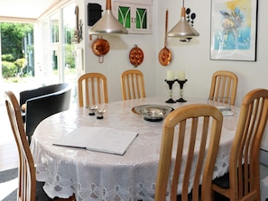 A dining area with a round table, six wooden chairs, a white lace tablecloth, a candle holder, and a painting on the wall.