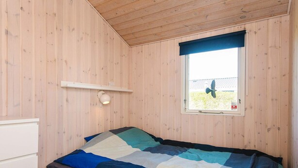 A bedroom with a wooden ceiling and walls, a bed with a multicolored quilt, a bedside table, and a window with a view of a bird flying outside.