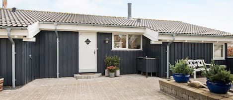 A residential building with a tiled roof, a dark facade, a white door, and a paved courtyard with potted plants.