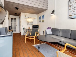 Una sala moderna con un sofá gris, una silla de madera, una mesita y una lámpara. Se ve una puerta que conduce a otra habitación y una pared con cuadros enmarcados.