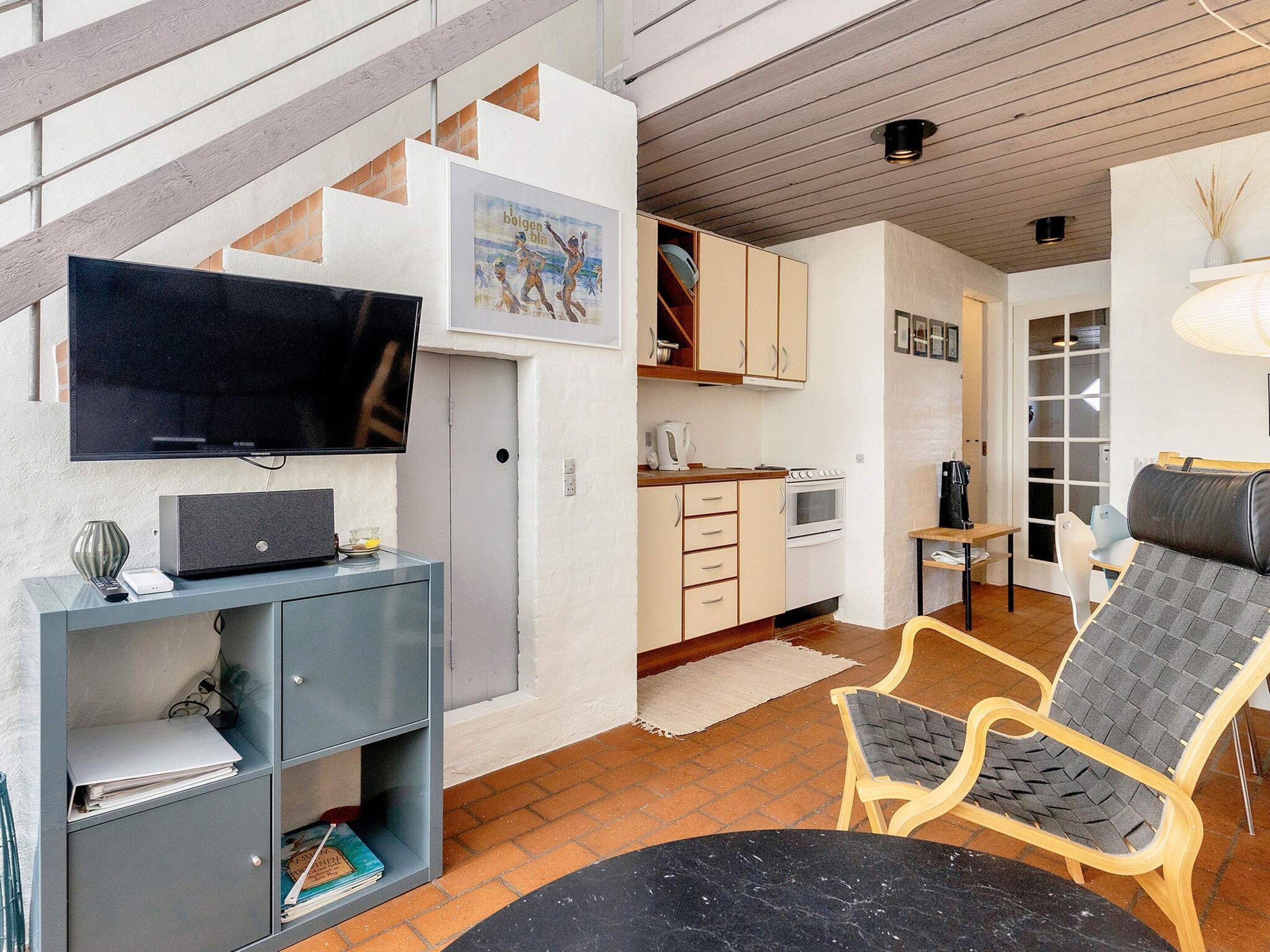 A modern living room with a flat-screen TV, a bookshelf, a dining area with a table and chairs, and a kitchen with cabinets and appliances.