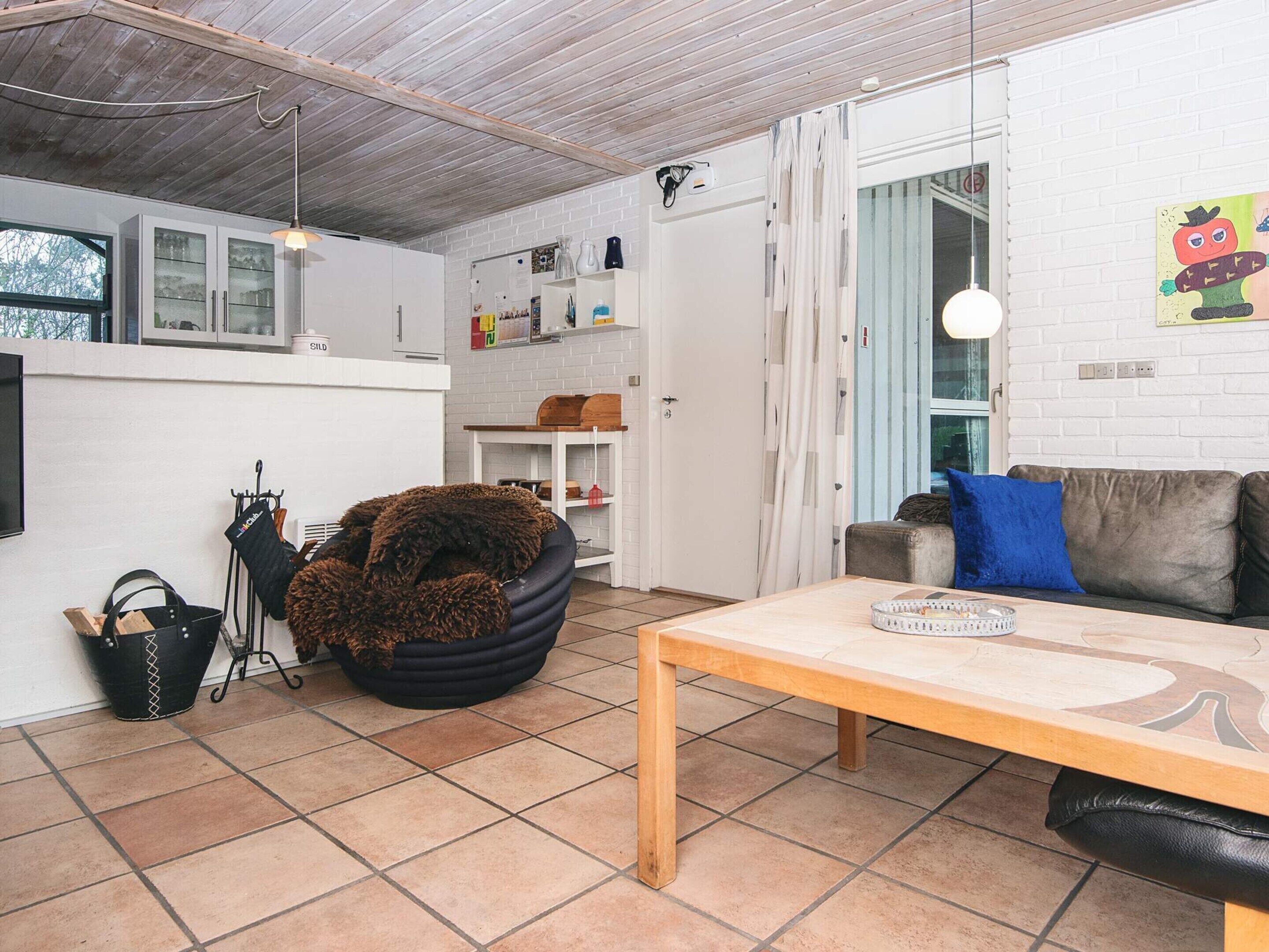 Una sala de estar acogedora con un sofá, una mesa de centro y un sillón negro y mullido. Se observa una zona de cocina con armarios y una ventana.