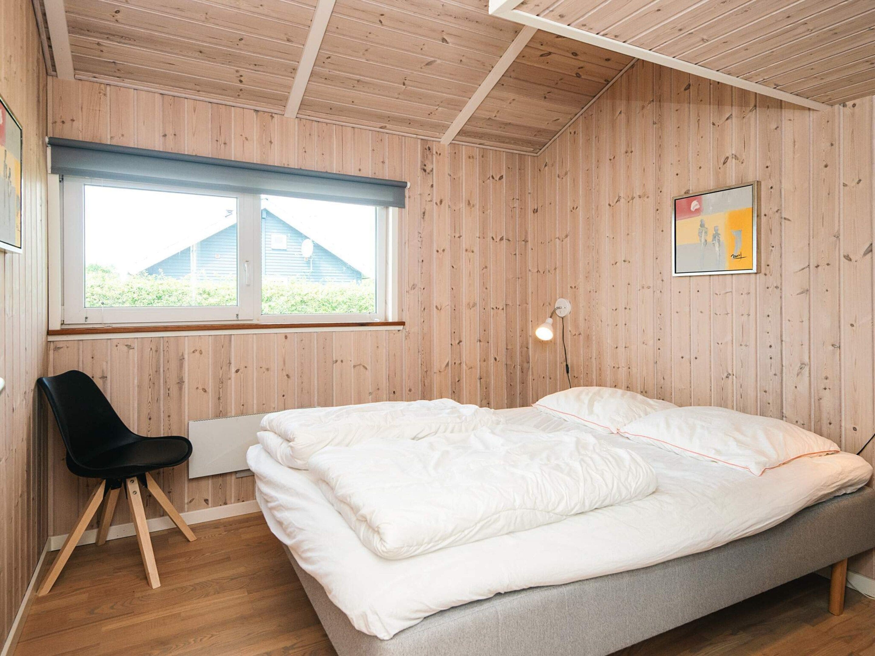 A bedroom with a bed, a chair, a window, and a wall-mounted light fixture.