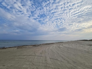 A beach with visible tire tracks on the sand and a clear sky with clouds.