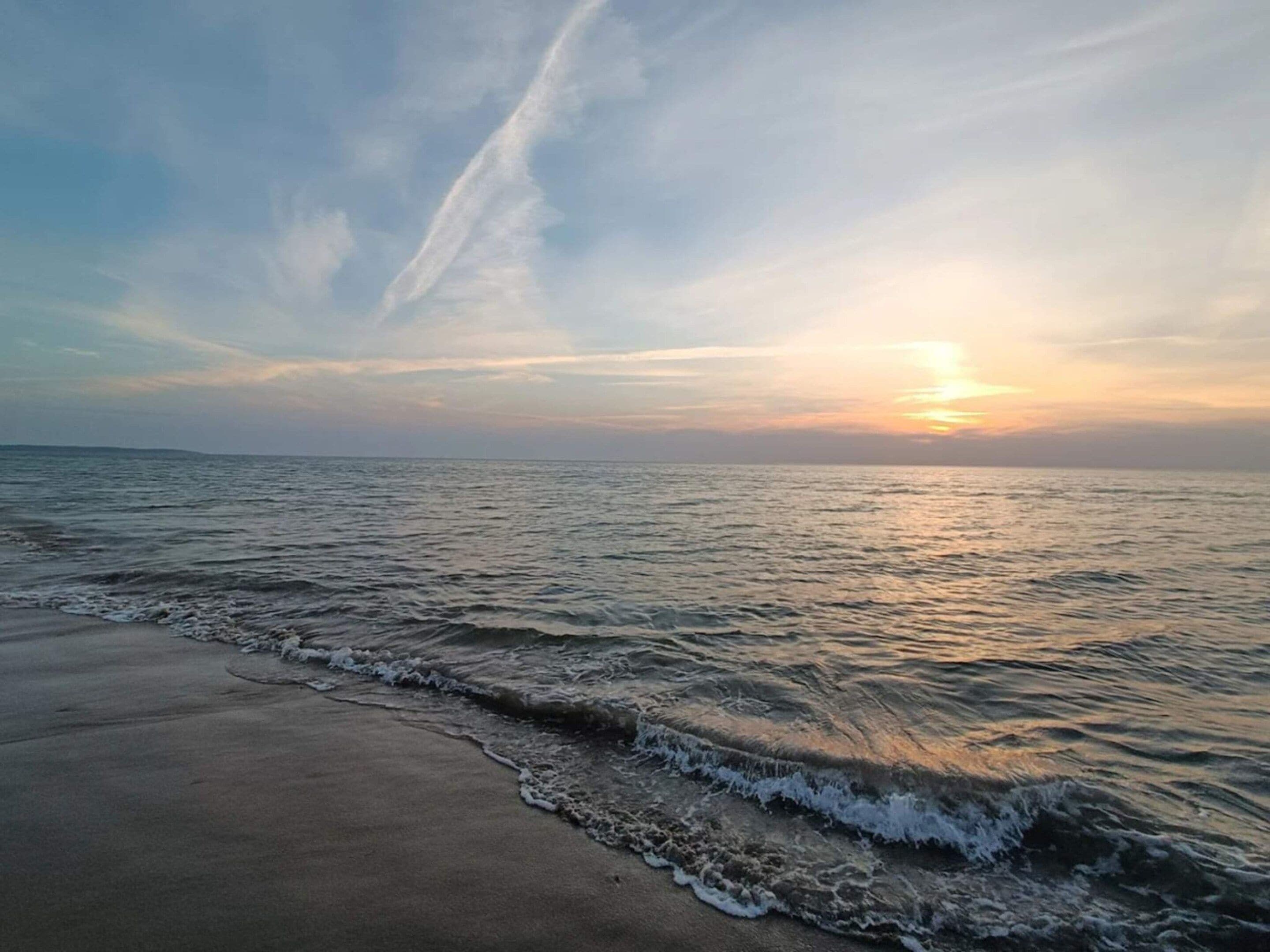 Una playa con olas suaves lamiendo la orilla, cielo despejado y un atardecer a lo lejos.