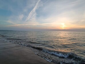 A beach with waves gently lapping onto the shore, a clear sky, and a sunset in the distance.