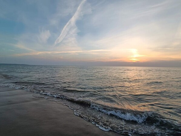 A beach with waves gently lapping onto the shore, a clear sky, and a sunset in the distance.