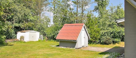 A small, single-story shed with a red roof, surrounded by a grassy yard and dense trees.