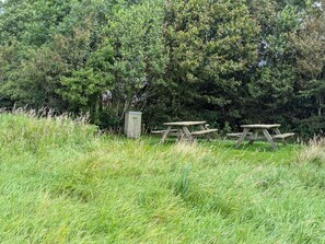 A picnic area with wooden benches and a small shed in a grassy field with dense trees in the background. - 6 Person Holiday Home in Oksbol (Oksbol)