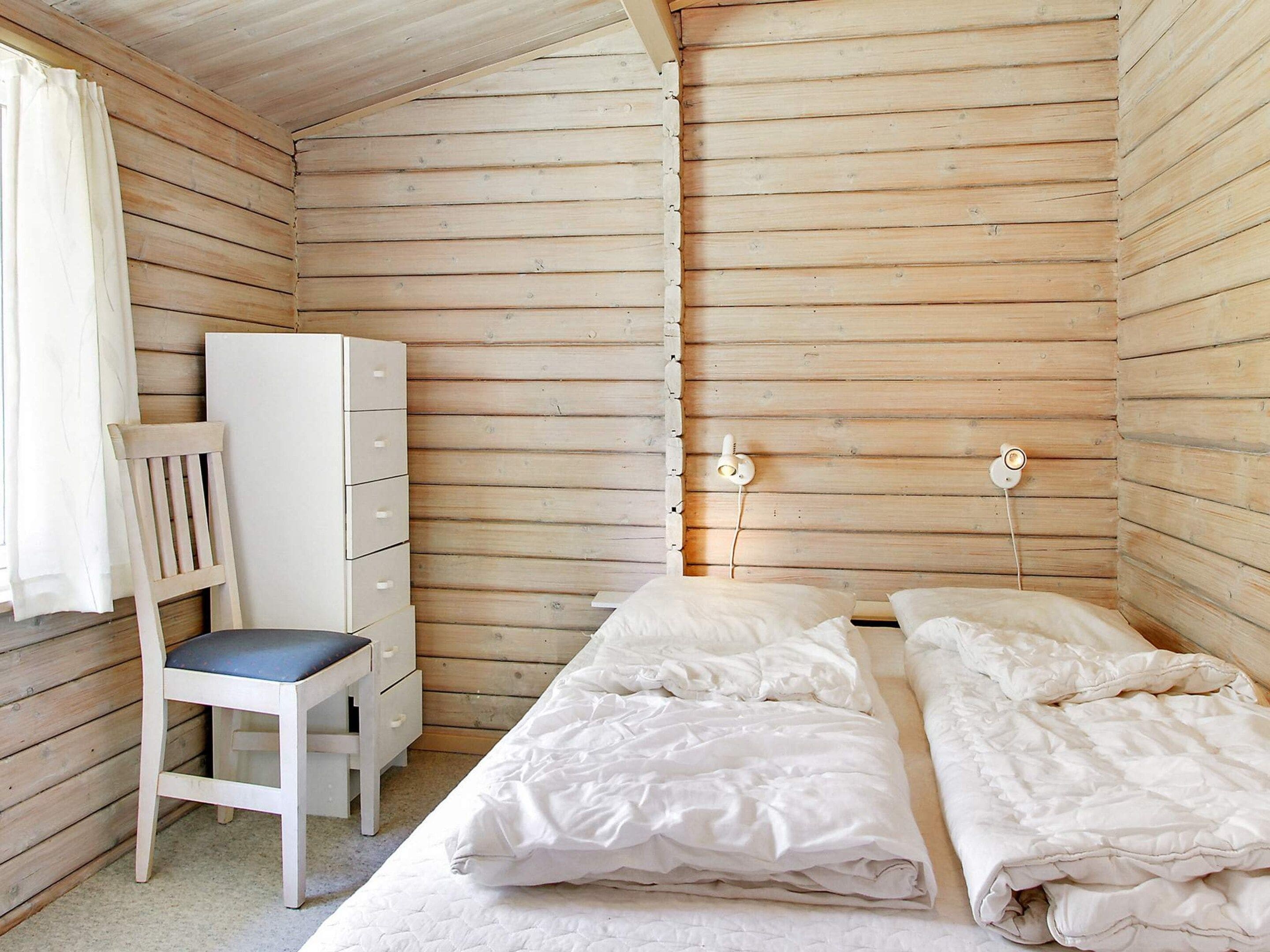 Quarto pequeno e acolhedor com paredes de madeira, uma cama de solteiro com roupa de cama branca, uma cadeira de madeira com almofada e um aparador branco.