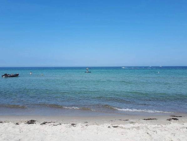 Une plage aux eaux calmes, un rivage sablonneux et quelques bateaux visibles au loin.