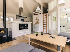 A modern interior with a central table, grey sofas, a black wood-burning stove, and a white kitchen area. - 4 Star Holiday Home in Vaeggerlose (Vaeggerlose)