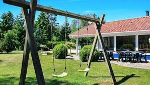 Un ensemble de balançoires en bois installé dans un espace herbeux, avec une terrasse couverte et des sièges d’extérieur en arrière-plan.