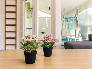 A room with a wooden table, two potted plants with flowers, a window with a view of a blue sky, and a ladder leaning against the wall.