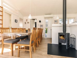 A dining area with wooden chairs and a glass table, a black stove with a chimney, and a white ceiling with wooden beams. - 4 Star Holiday Home in Vaeggerlose (Vaeggerlose)