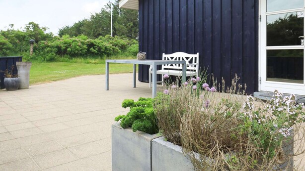 A patio with a table and a white chair, a concrete planter with greenery, and a dark blue building with a white window. - 8 Person Holiday Home in Hjorring (Hjørring)