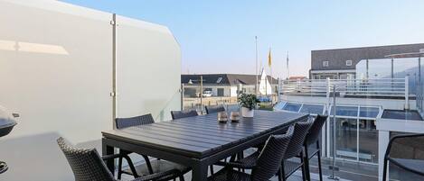 A rooftop terrace with a table and chairs, a potted plant, and a view of a sailboat and buildings in the background.
