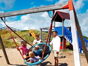 A playground with a swing set and a slide, surrounded by a sandy area and grassy dunes. - 5 Star Holiday Home in Ringkobing (Ringkobing)