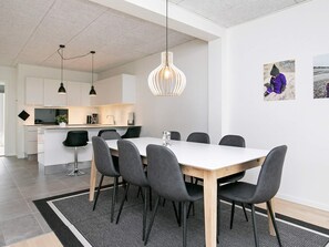 A modern dining area with a white table and black chairs, a kitchen in the background, and a hanging light fixture.