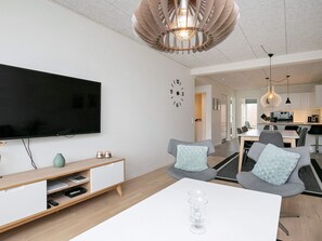 Ein modernes Wohnzimmer mit einem Flachbildfernseher, einem Couchtisch und einem Essbereich, wobei im Hintergrund eine Küche zu sehen ist.