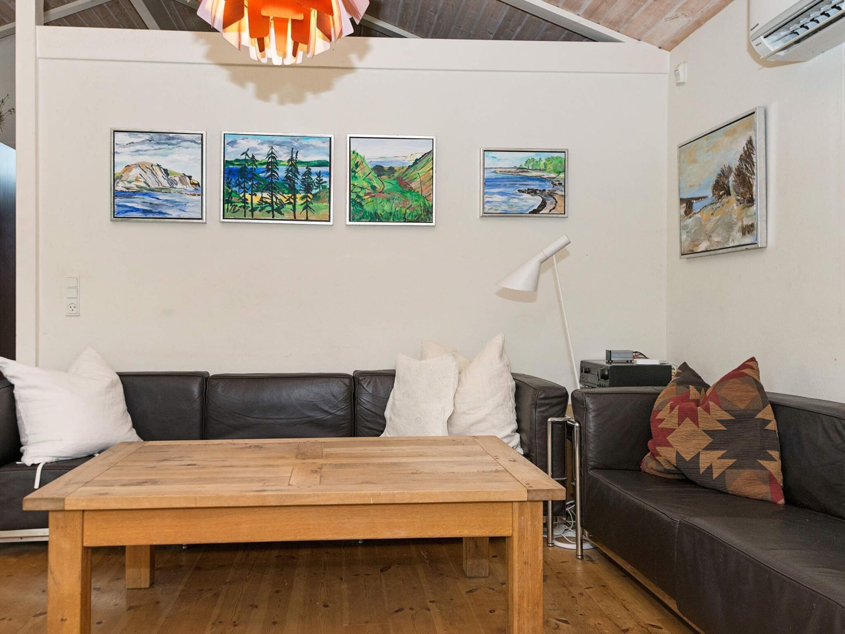 Sala de estar com mesa de centro de madeira, sofá de couro preto e luminária de chão branca. Quatro quadros emoldurados decoram a parede.