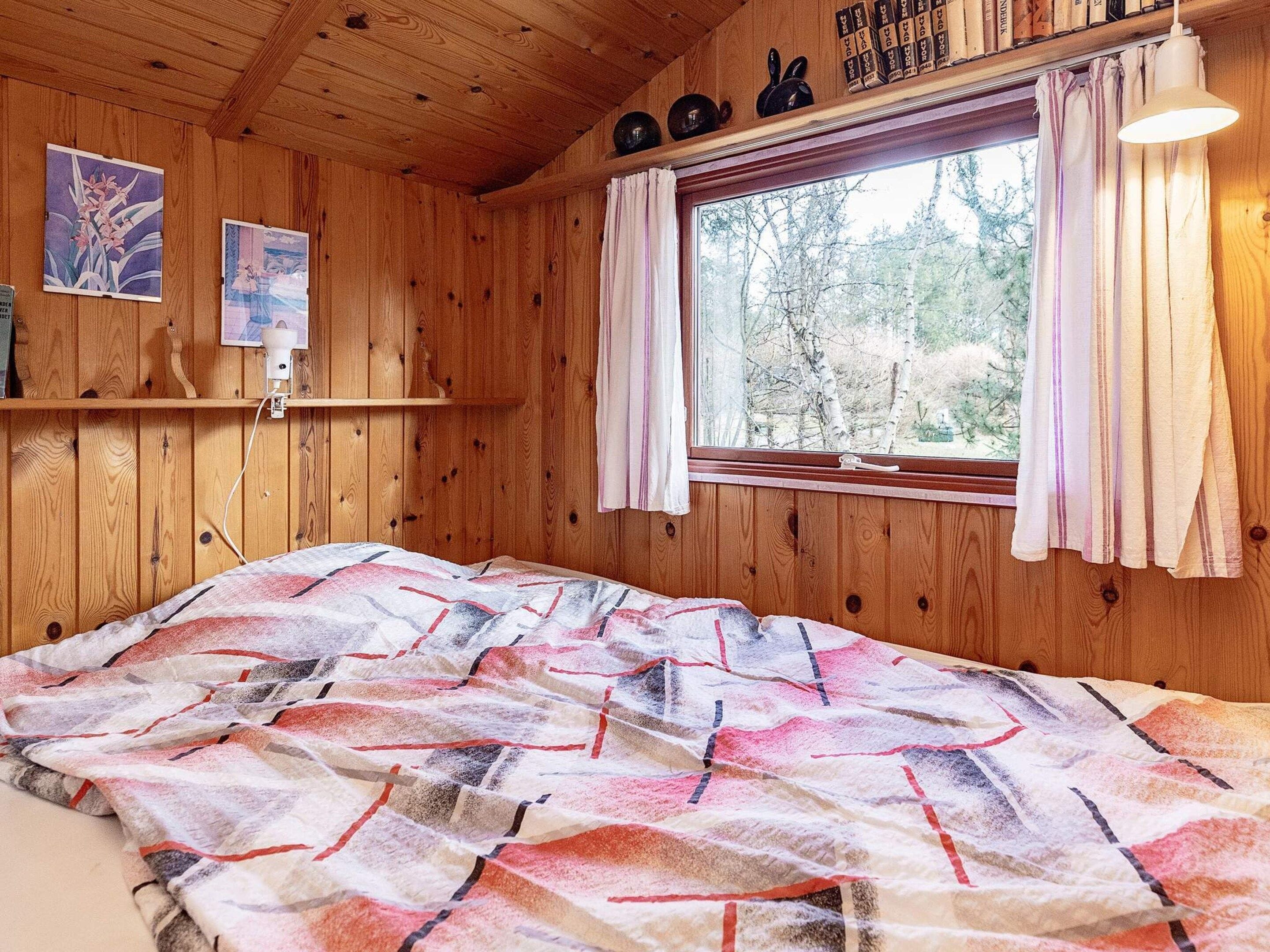 Una habitación de madera con una cama, una ventana con cortinas, y una estantería con libros y objetos decorativos.
