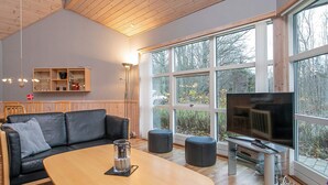 A modern living room with a wooden ceiling, a black leather sofa, a glass coffee table, and a large window offering a view of trees and a building. - 4 Star Holiday Home in Laeso (Læsø)