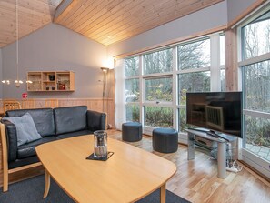 Ett modernt vardagsrum med ett träpanelat tak, en svart lädersoffa, ett soffbord i glas och ett stort fönster med utsikt över träd och en byggnad.