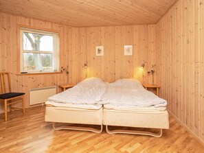 A room with wooden walls and floors, a bed with two mattresses, a chair, a window with a view of trees, and two wall-mounted lamps.